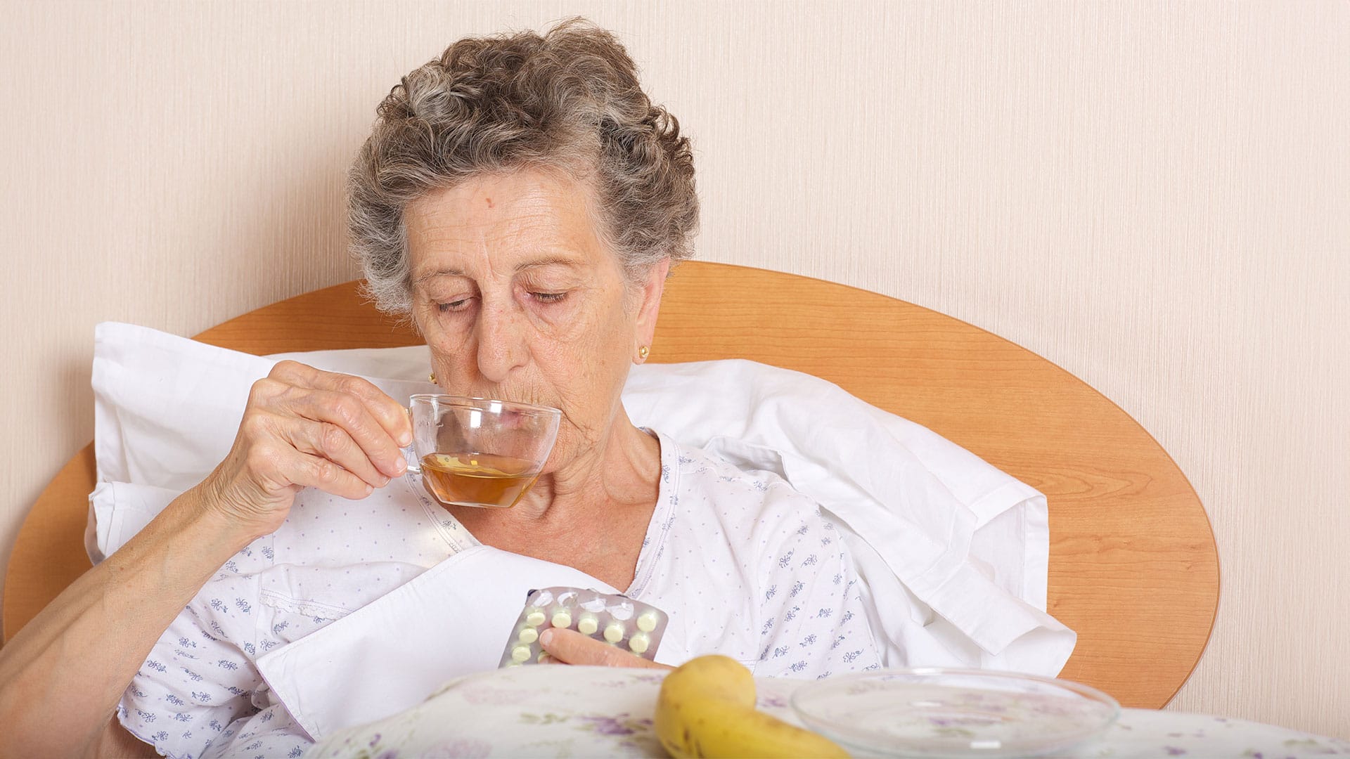 Image of an older woman sitting on a cozy chair, holding a steaming cup of tea while battling the flu, wrapped in a blanket.